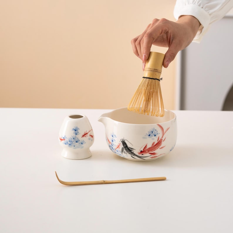 Pu&ograve; includere: Una ciotola di matcha in ceramica bianca con un design di pesci blu e rossi, un porta-frusta per matcha in ceramica bianca con un design floreale blu e rosso, una frusta per matcha in bamb&ugrave; e una paletta per matcha in bamb&ugrave;.