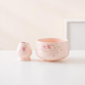 May include: A light pink ceramic tea bowl and a small vase. Both are decorated with pink cherry blossom branches. The bowl is wide and shallow, while the vase is a small, rounded shape. The background is a soft, neutral color.