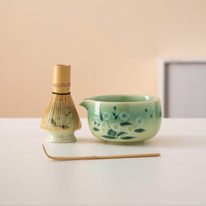 Op de afbeelding: Een groene keramische matcha-kom met een bloemenpatroon en een bamboe matcha-garde. De kom heeft een tuit en staat op een wit oppervlak. Er ligt ook een bamboe-garde op het oppervlak.