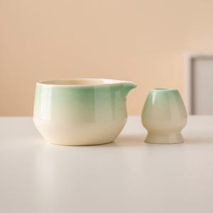 May include: A ceramic matcha tea set featuring a cream-colored bowl with a spout and a small, rounded whisk holder. Both pieces have a gradient of green at the top, transitioning to cream. The set is designed for preparing matcha tea.