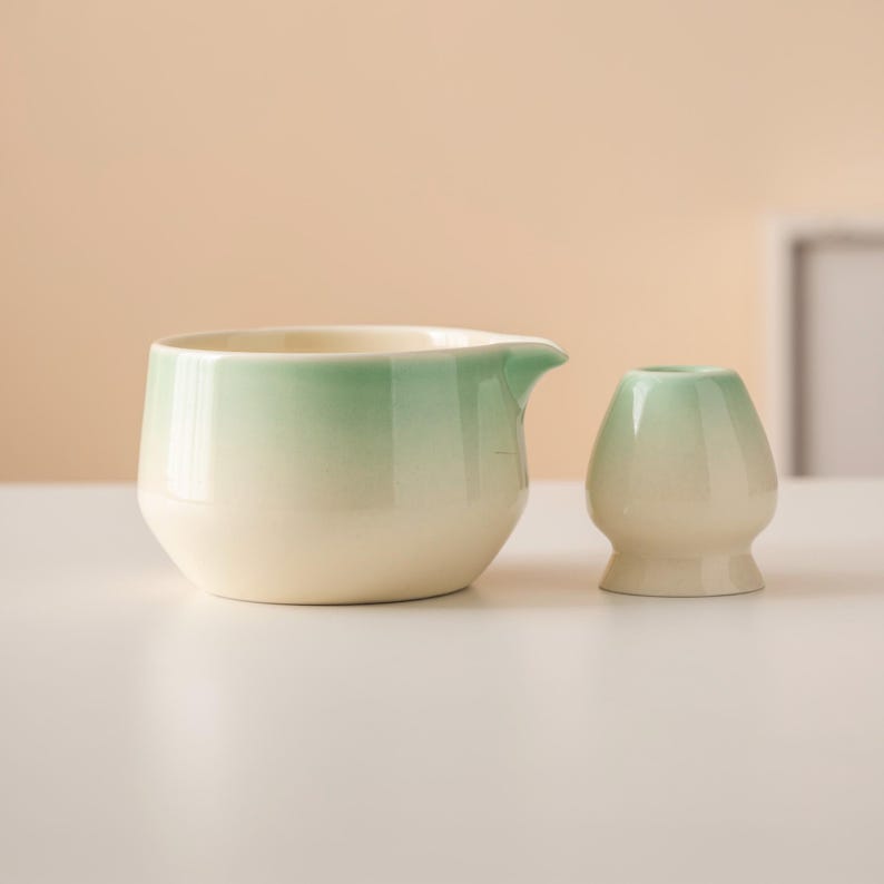 May include: A ceramic matcha tea set featuring a cream-colored bowl with a spout and a small, rounded whisk holder. Both pieces have a gradient of green at the top, transitioning to cream. The set is designed for preparing matcha tea.