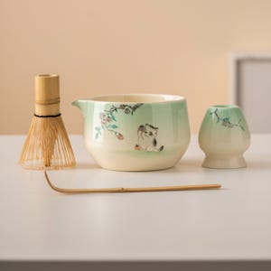 May include: A matcha tea set featuring a bamboo whisk, a ceramic bowl with a spout, a small ceramic holder, and a bamboo scoop. The bowl and holder are decorated with a light green glaze and a cat illustration.