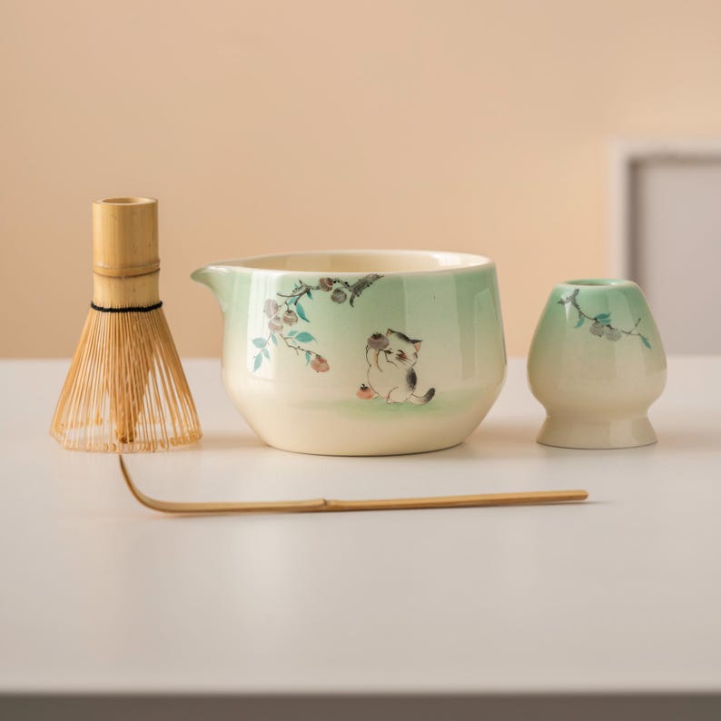May include: A matcha tea set featuring a bamboo whisk, a ceramic bowl with a spout, a small ceramic holder, and a bamboo scoop. The bowl and holder are decorated with a light green glaze and a cat illustration.