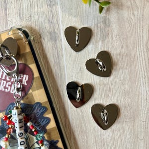 May include: Four silver heart-shaped charms with a loop on the back, next to a phone case with a keychain. The keychain has a beaded design with the word "GOODGIRL" spelled out. The background is a light wood grain.