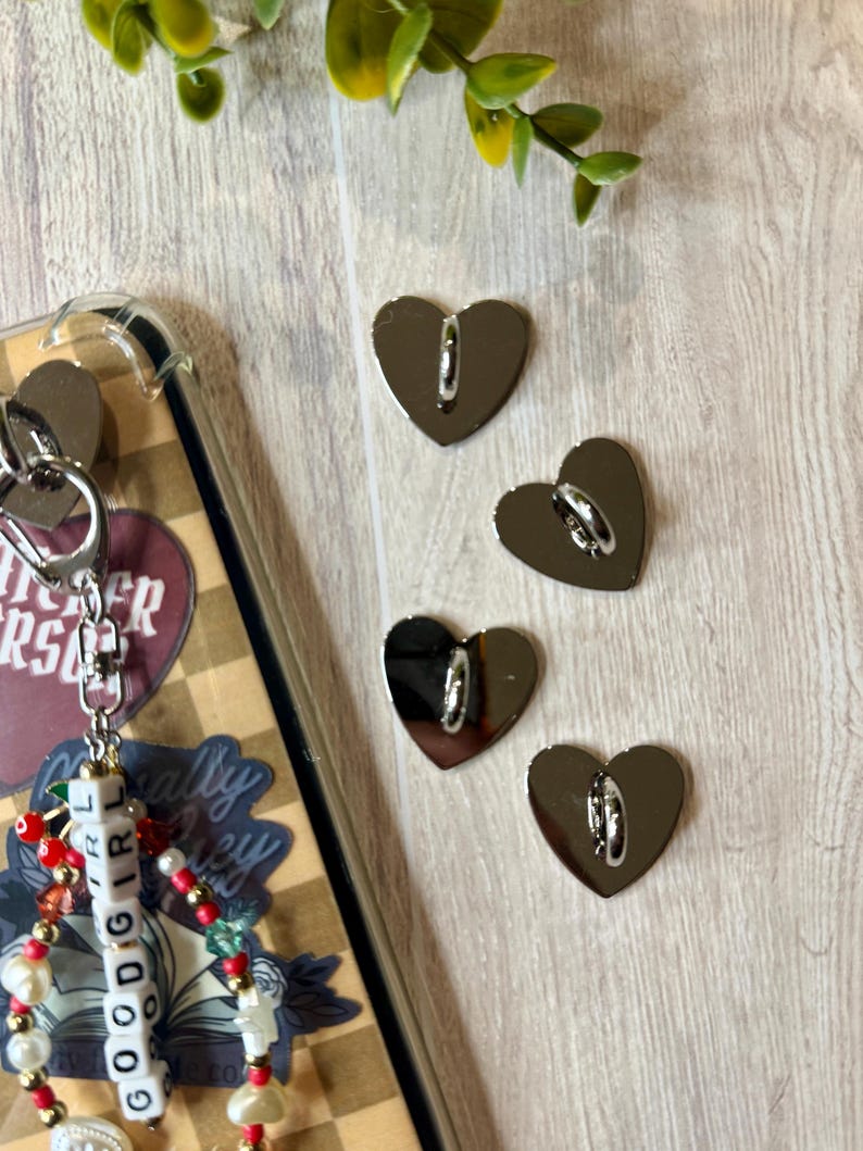 May include: Four silver heart-shaped charms with a loop on the back, next to a phone case with a keychain. The keychain has a beaded design with the word "GOODGIRL" spelled out. The background is a light wood grain.