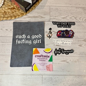 Puede incluir: Una colección de artículos que incluye una pancarta de fieltro gris con el texto "such a good fucking girl" en blanco, un paquete de mezcla para bebidas Craftmix Passionfruit Paloma y varias pegatinas.