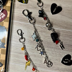May include: Three keychains with silver clasps and decorative charms. Charms include a lightning bolt, stars, a heart, and a microphone. The keychains are on a light wood-grain surface with heart-shaped phone grips.