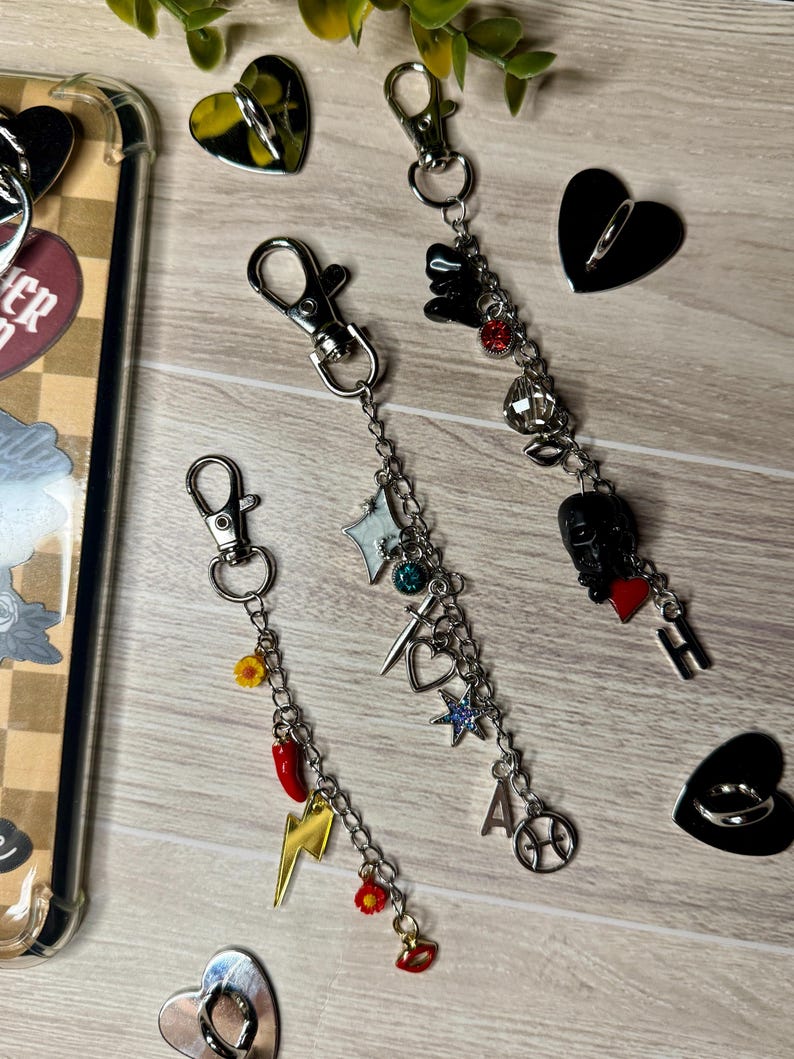May include: Three keychains with silver clasps and decorative charms. Charms include a lightning bolt, stars, a heart, and a microphone. The keychains are on a light wood-grain surface with heart-shaped phone grips.