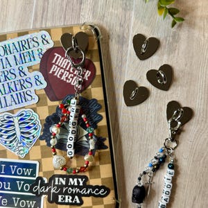 May include: A clear phone case with stickers and two keychains. One keychain has a beaded bracelet with the word "GOODGIRL". The other keychain has a black skull charm and the word "BADDIE". Three heart-shaped metal clasps are also visible.