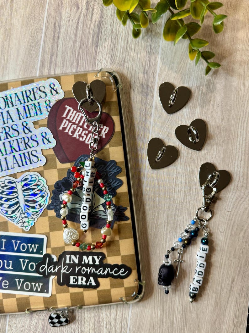May include: A clear phone case with stickers and two keychains. One keychain has a beaded bracelet with the word "GOODGIRL". The other keychain has a black skull charm and the word "BADDIE". Three heart-shaped metal clasps are also visible.