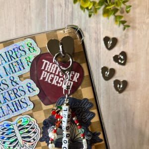 May include: A silver keychain with a heart-shaped charm and a beaded strand spelling "GOOD GIRL." The keychain is attached to a clear phone case with stickers. Additional silver heart charms are scattered nearby. The background has a checkered pattern.