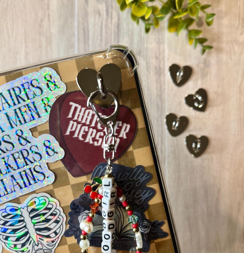May include: A silver keychain with a heart-shaped charm and a beaded strand spelling "GOOD GIRL." The keychain is attached to a clear phone case with stickers. Additional silver heart charms are scattered nearby. The background has a checkered pattern.