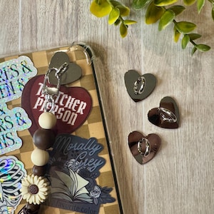 May include: A clear phone case with a checkerboard pattern and decorative stickers. A beaded charm with wooden beads and a flower is attached. Three silver heart-shaped charms with rings are also visible.