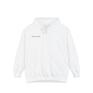 Puede incluir: Una sudadera con capucha blanca con un bolsillo delantero y una capucha con cordón. El texto "Forever Lucky" está impreso en una fuente pequeña y negra en la parte superior del pecho. La sudadera está hecha de un material suave y cómodo.