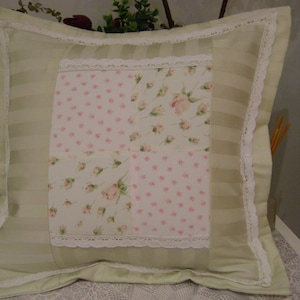 May include: A green and white pillow with a patchwork design featuring pink floral patterns. The pillow has a white lace trim around the edges.