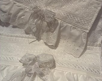 May include: White, eyelet-trimmed bedding set featuring intricate lace details and delicate ribbon bows. The set includes a top sheet and a decorative element with a ruffled edge. The design is classic and elegant, suitable for a bedroom.