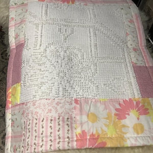 May include: A patchwork quilt with a white, textured center and floral patterned borders. The quilt features pink, yellow, and white daisy and floral prints, with a pink and white striped pattern. The quilt is a mix of textures and colors.