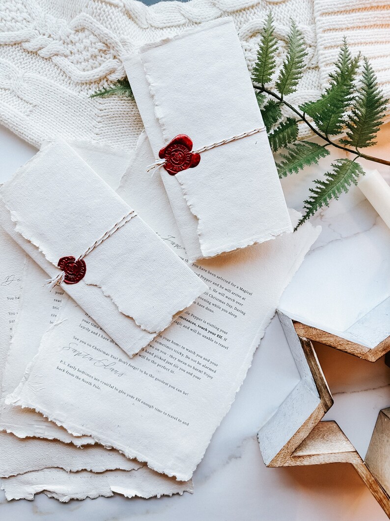 May include: Handmade paper letters with torn edges, sealed with red wax stamps and tied with twine. The letters have handwritten text and are arranged with a white knitted sweater and a wooden star.