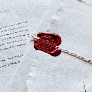 May include: Close-up of stationery with a red wax seal. The seal is on a white paper with torn edges. The paper has handwritten text. The seal is attached to the paper with a white and gold string. The words "FROM THE DESK OF SANTA OFFICIAL" are visible.