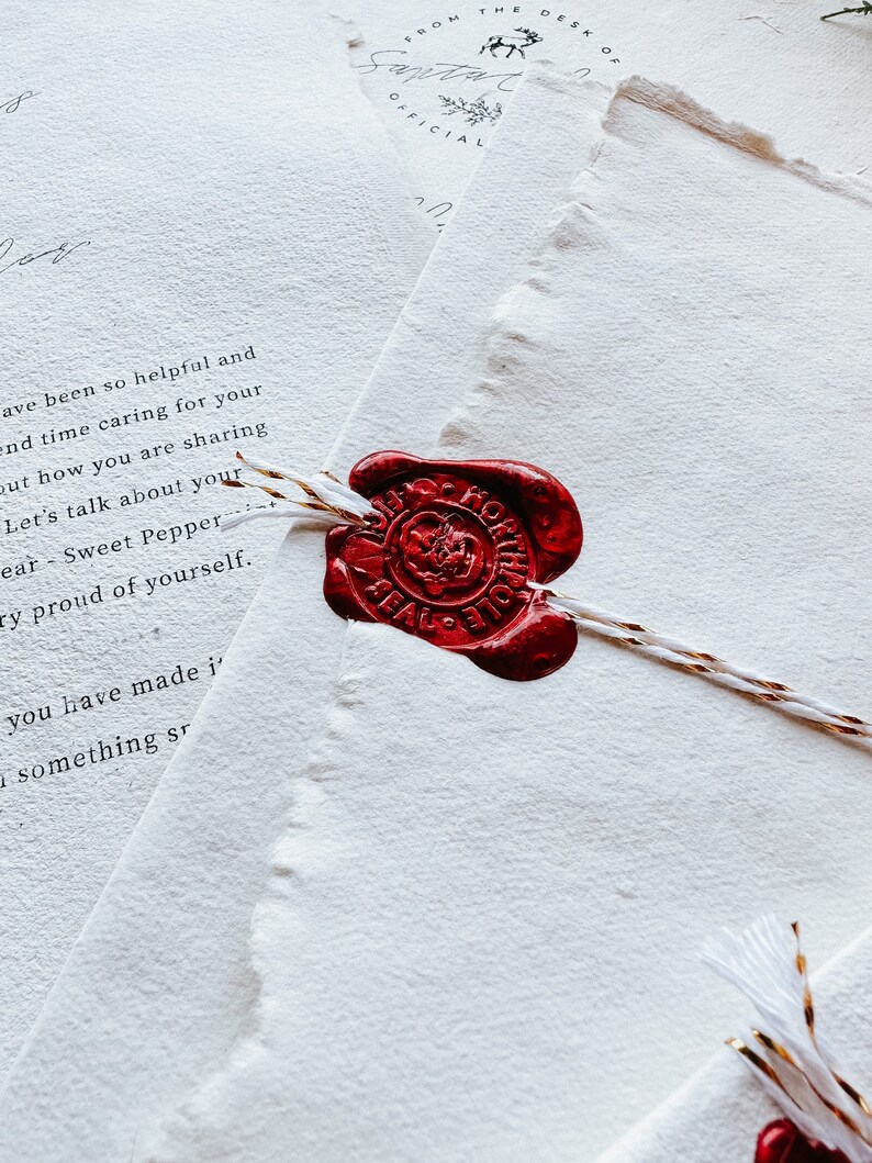 May include: Close-up of stationery with a red wax seal. The seal is on a white paper with torn edges. The paper has handwritten text. The seal is attached to the paper with a white and gold string. The words "FROM THE DESK OF SANTA OFFICIAL" are visible.