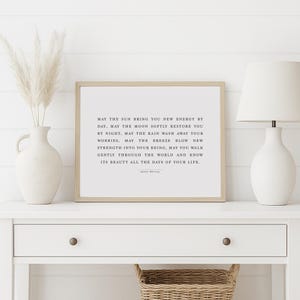 May include: A framed print with a light brown frame and a white background. The print features a quote about finding strength and beauty in life. The quote reads: "May the sun bring you new energy by day, may the moon softly restore you by night, may the rain wash away your worries, may the breeze blow new strength into your being, may you walk gently through the world and know its beauty all the days of your life."