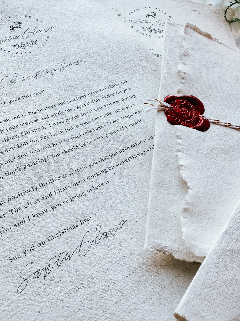 May include: Close-up of a stack of textured white paper letters, one sealed with a red wax stamp. The letters feature elegant cursive handwriting and the words "The Desk of Santa Claus" at the top.