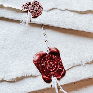 May include: Close-up of cream-colored paper with torn edges, sealed with red wax seals. The seals have intricate designs and are tied with white and gold string. The paper is layered, creating a textured effect.