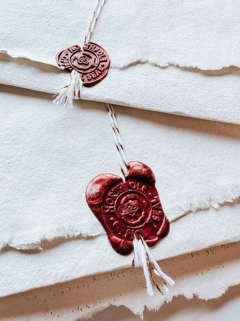 May include: Close-up of cream-colored paper with torn edges, sealed with red wax seals. The seals have intricate designs and are tied with white and gold string. The paper is layered, creating a textured effect.