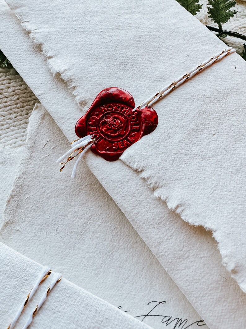 May include: Close-up of cream-colored stationery with a torn edge, sealed with a red wax seal. The seal reads "NORTH POLE SEAL" and is tied with a white and gold string. The stationery has a textured surface.