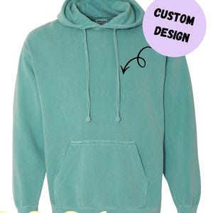 以下が含まれることがあります： ドローコード付きフードとカンガルーポケットが付いた、ティール色のComfort Colorsのパーカーです。「CUSTOM DESIGN」と矢印が描かれた円形のグラフィックが含まれています。「Fast Shipping」の文字も表示されています。