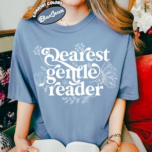 May include: A blue t-shirt with the words "Dearest Gentle Reader" in white script and floral accents. The shirt is part of the Comfort Colors collection and is labeled "Blue Jean". The person is holding an open book.