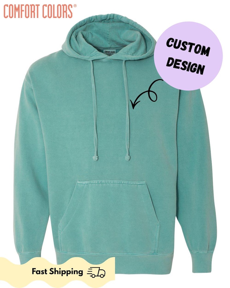 以下が含まれることがあります： ドローコード付きフードとカンガルーポケットが付いた、ティール色のComfort Colorsのパーカーです。「CUSTOM DESIGN」と矢印が描かれた円形のグラフィックが含まれています。「Fast Shipping」の文字も表示されています。