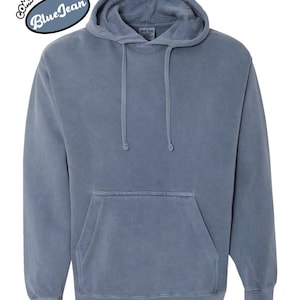 以下が含まれることがあります： フロントポケットとドローストリングフードが付いた、ブルージーンズカラーのパーカー。「Comfort Colors Blue Jean」の文字が左上に円形デザインで配置されています。