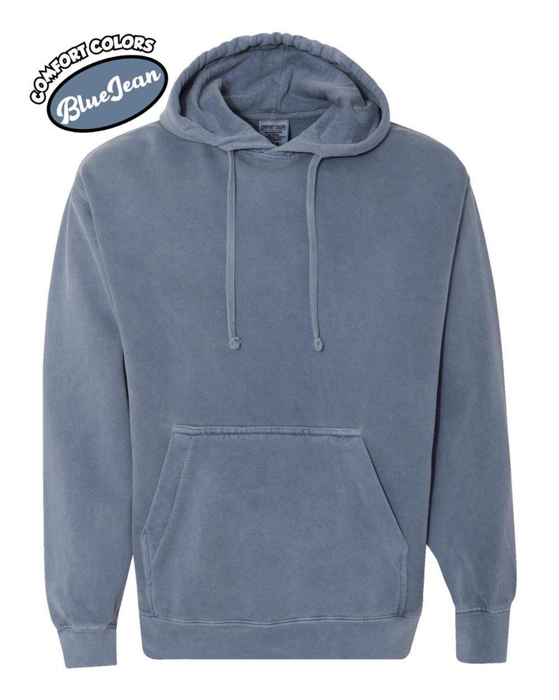 以下が含まれることがあります： フロントポケットとドローストリングフードが付いた、ブルージーンズカラーのパーカー。「Comfort Colors Blue Jean」の文字が左上に円形デザインで配置されています。