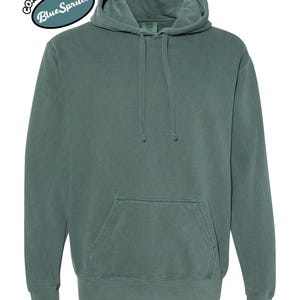 以下が含まれることがあります： ドローストリングフードとカンガルーポケットが付いた、ブルースプルース色のパーカー。「COMFORT COLORS」と「Blue Spruce」の文字が円形にデザインされています。