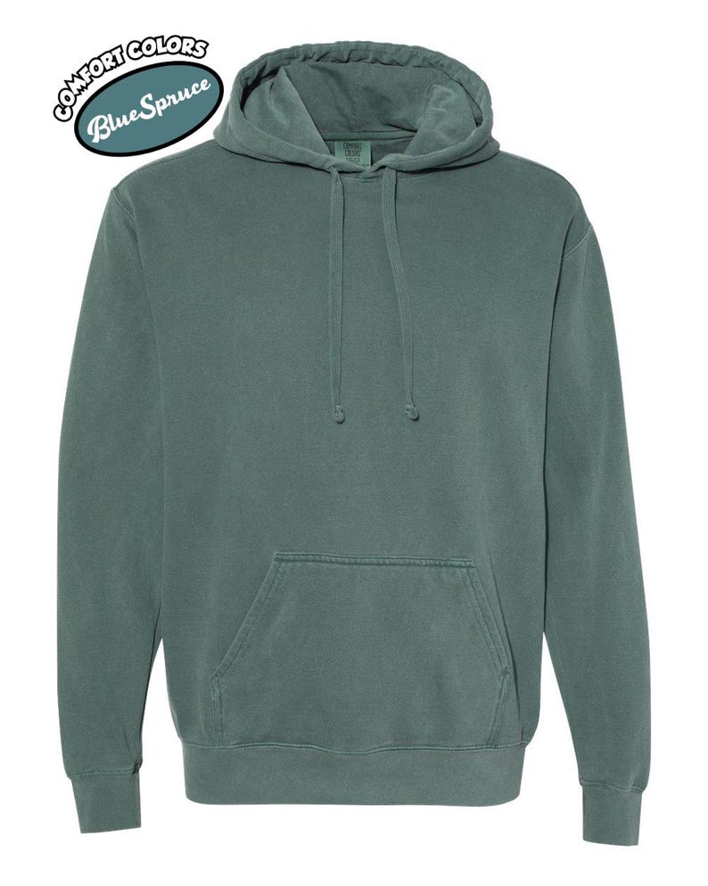 以下が含まれることがあります： ドローストリングフードとカンガルーポケットが付いた、ブルースプルース色のパーカー。「COMFORT COLORS」と「Blue Spruce」の文字が円形にデザインされています。