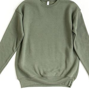 May include: A solid olive green crewneck sweatshirt. The long-sleeved top has a ribbed hem and cuffs, and a crew neckline. The sweatshirt is made of a soft, comfortable material.