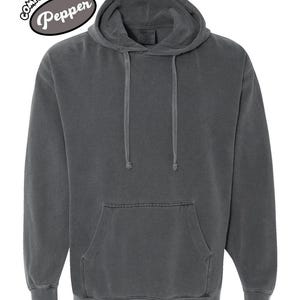 以下が含まれることがあります： ダークグレーのComfort Colors Pepperのパーカー。フードにはドローコード、カンガルーポケットが付いています。パーカーには、左上に円形のデザインでComfort Colorsのロゴが配置されています。