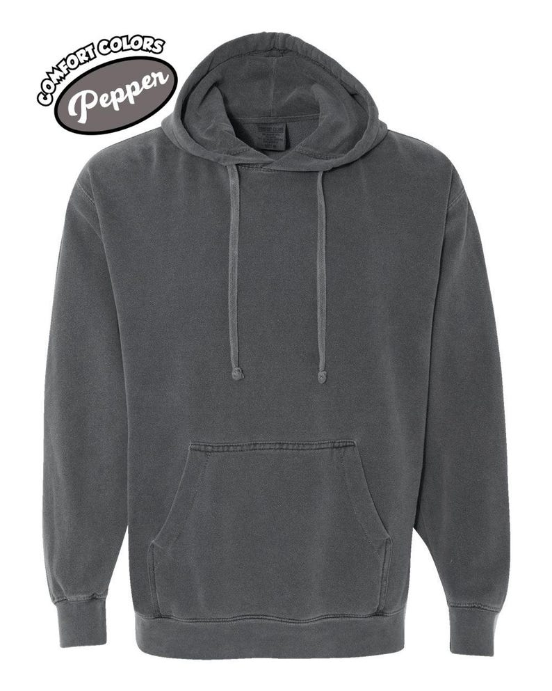 以下が含まれることがあります： ダークグレーのComfort Colors Pepperのパーカー。フードにはドローコード、カンガルーポケットが付いています。パーカーには、左上に円形のデザインでComfort Colorsのロゴが配置されています。