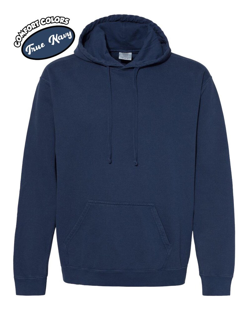 以下が含まれることがあります： フロントポケットとドローストリングフードが付いたネイビーブルーのパーカー。パーカーの左上に白い楕円で「Comfort Colors True Navy」の文字が入っています。この衣服は柔らかく快適な素材で作られています。