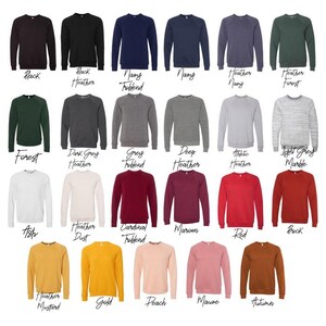 May include: A collection of crewneck sweatshirts in various colors, including black, navy, grey, red, and gold. Each sweatshirt has a solid color and the name of the color is printed below. The sweatshirts are made of a soft material.
