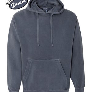 以下が含まれることがあります： デニムカラーのフード付きパーカー。フロントポケットとドローストリングフードが付いています。「Comfort Colors」と「Denim」の文字が白と黒の楕円形のパッチにプリントされています。柔らかく快適な素材で作られています。