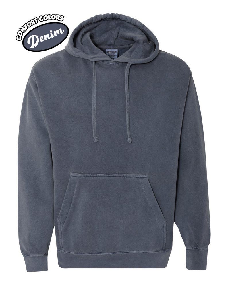 以下が含まれることがあります： デニムカラーのフード付きパーカー。フロントポケットとドローストリングフードが付いています。「Comfort Colors」と「Denim」の文字が白と黒の楕円形のパッチにプリントされています。柔らかく快適な素材で作られています。