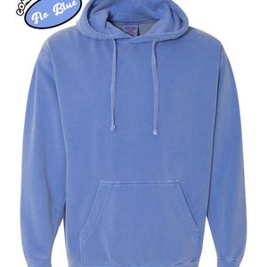 以下が含まれることがあります： カンガルーポケットとドローストリングフードが付いた青いパーカー。パーカーは単色で、「Comfort Colors Flo Blue」の文字が円形にデザインされています。