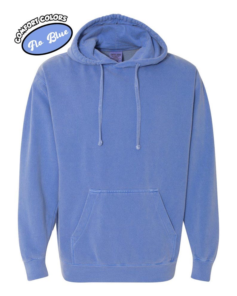 以下が含まれることがあります： カンガルーポケットとドローストリングフードが付いた青いパーカー。パーカーは単色で、「Comfort Colors Flo Blue」の文字が円形にデザインされています。