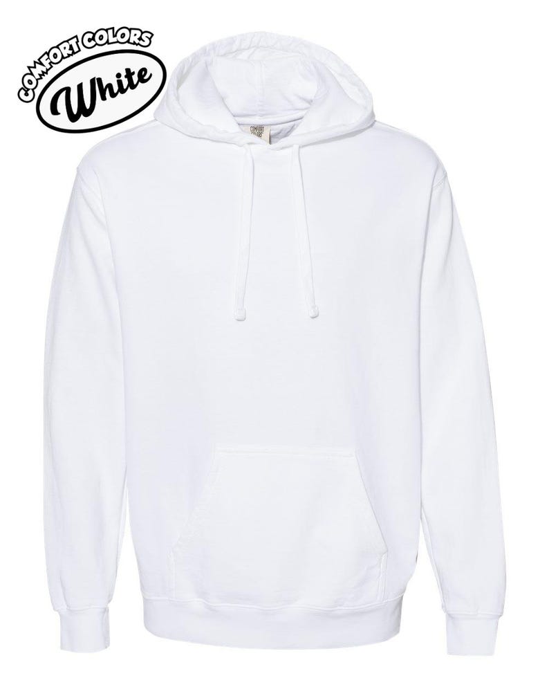 以下が含まれることがあります： フロントポケットとドローストリングフードが付いた白いパーカー。「Comfort Colors White」の文字が左上の白黒の楕円の中にあります。パーカーは柔らかく快適な素材で作られています。