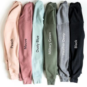 May include: A collection of six sweatshirts in various colors, including peach, mauve, dusty blue, military green, heather deep, and black. Each sweatshirt is folded, with the color name printed vertically on the sleeve. The sweatshirts are displayed against a white background.