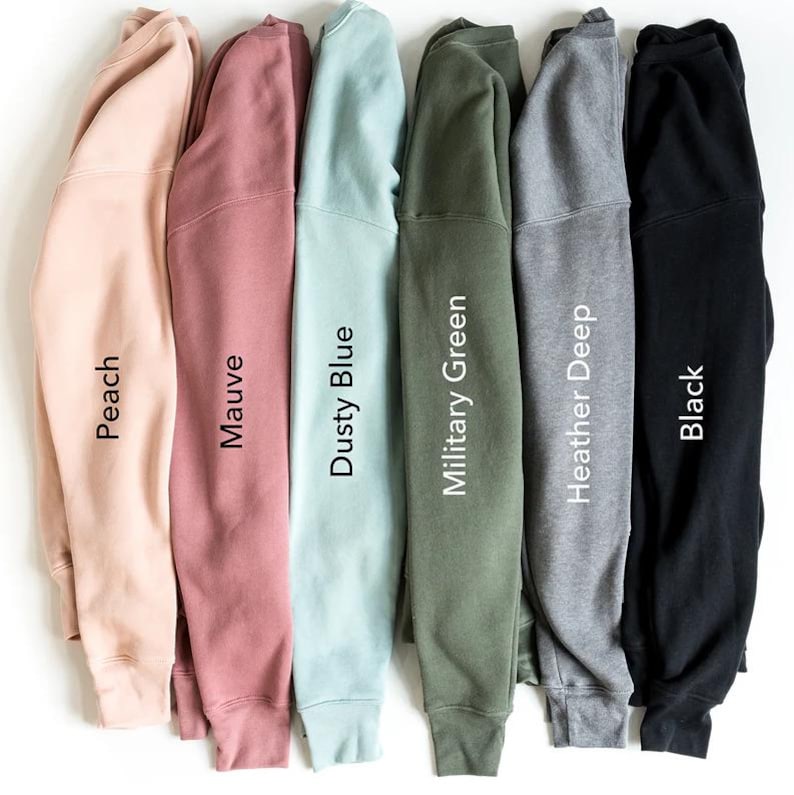 May include: A collection of six sweatshirts in various colors, including peach, mauve, dusty blue, military green, heather deep, and black. Each sweatshirt is folded, with the color name printed vertically on the sleeve. The sweatshirts are displayed against a white background.