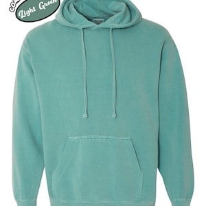 以下が含まれることがあります： ドローコード付きフードとカンガルーポケットが付いた、ライトグリーンのパーカー。スウェットシャツは単色で、「Comfort Colors Light Green」の文字が円形にデザインされています。