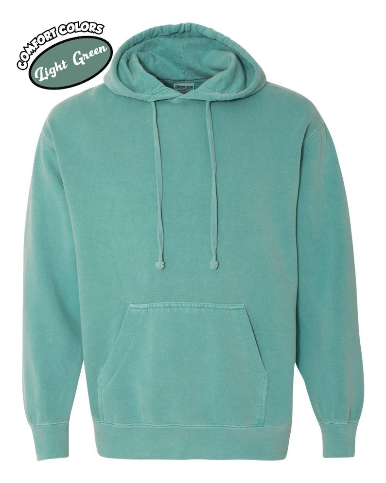 以下が含まれることがあります： ドローコード付きフードとカンガルーポケットが付いた、ライトグリーンのパーカー。スウェットシャツは単色で、「Comfort Colors Light Green」の文字が円形にデザインされています。